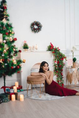 Portrait of pretty young girl cozy sit down on the floor, smiling wear red gown in decorated Christmas living room indoors
