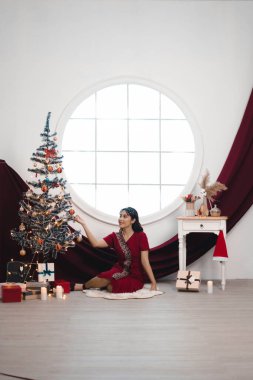 Portrait of pretty young girl cozy sit down on the floor, smiling wear red gown in decorated Christmas living room indoors