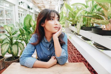 Thoughtful Asian woman keeps hand on chin looks pensively above dressed in blue shirt poses thinks about future