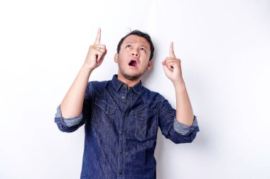 Shocked Asian man wearing blue shirt pointing at the copy space on top of him, isolated by white background