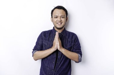 Smiling young Asian man wearing blue shirt, gesturing traditional greeting isolated over white background