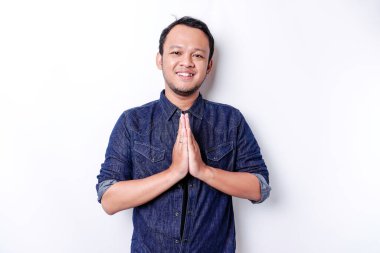 Smiling young Asian man wearing blue shirt, gesturing traditional greeting isolated over white background