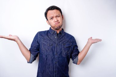 A portrait of an Asian man wearing a blue shirt looks so confused between choices, isolated by a white background