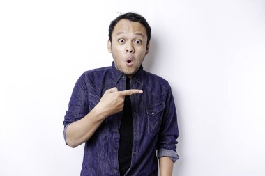 Shocked Asian man wearing blue shirt pointing at the copy space beside him, isolated by white background