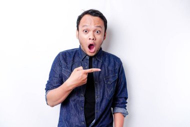 Shocked Asian man wearing blue shirt pointing at the copy space beside him, isolated by white background