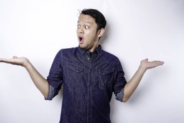 Shocked Asian man wearing blue shirt pointing at the copy space beside him, isolated by white background