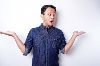 Shocked Asian man wearing blue shirt pointing at the copy space beside him, isolated by white background