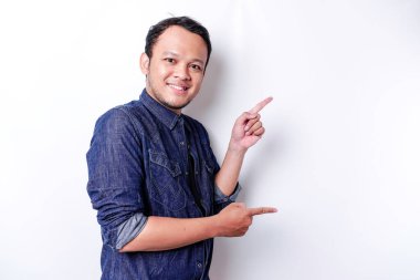 Excited Asian man wearing blue shirt pointing at the copy space beside him, isolated by white background