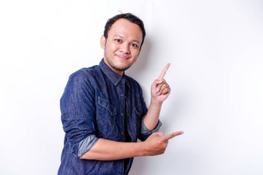 Excited Asian man wearing blue shirt pointing at the copy space beside him, isolated by white background