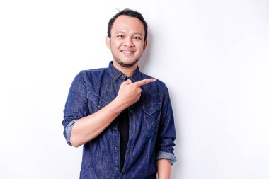 Excited Asian man wearing blue shirt pointing at the copy space beside him, isolated by white background