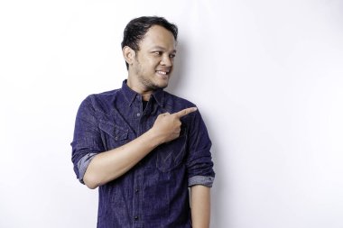 Excited Asian man wearing blue shirt pointing at the copy space beside him, isolated by white background