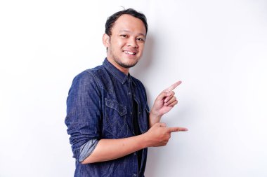 Excited Asian man wearing blue shirt pointing at the copy space beside him, isolated by white background