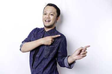 Excited Asian man wearing blue shirt pointing at the copy space beside him, isolated by white background