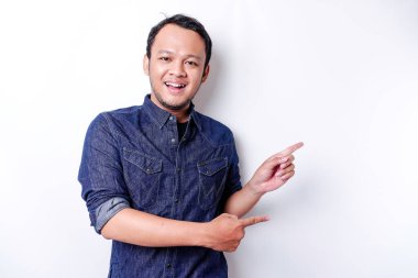 Excited Asian man wearing blue shirt pointing at the copy space beside him, isolated by white background