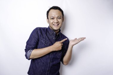 Excited Asian man wearing blue shirt pointing at the copy space beside him, isolated by white background