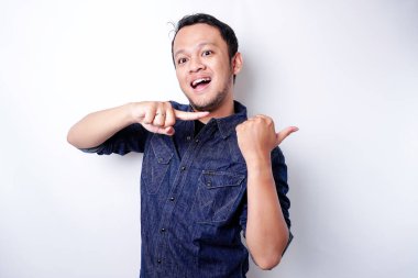 Excited Asian man wearing blue shirt pointing at the copy space beside him, isolated by white background