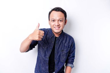 Excited Asian man wearing blue shirt gives thumbs up hand gesture of approval, isolated by white background