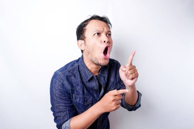 Shocked Asian man wearing blue shirt pointing at the copy space beside him, isolated by white background