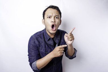 Shocked Asian man wearing blue shirt pointing at the copy space beside him, isolated by white background