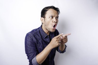Shocked Asian man wearing blue shirt pointing at the copy space beside him, isolated by white background