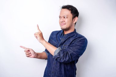 Excited Asian man wearing blue shirt pointing at the copy space beside him, isolated by white background