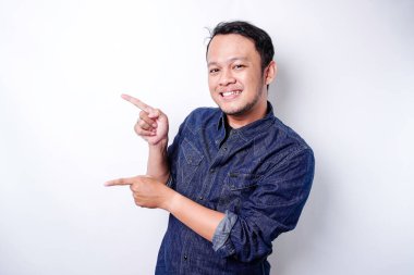 Excited Asian man wearing blue shirt pointing at the copy space beside him, isolated by white background