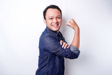 Excited Asian man wearing a blue shirt showing strong gesture by lifting his arms and muscles smiling proudly