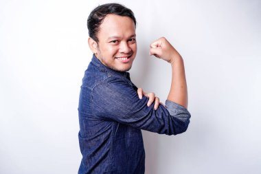 Excited Asian man wearing a blue shirt showing strong gesture by lifting his arms and muscles smiling proudly