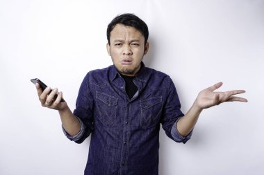 A thoughtful young Asian man is wearing blue shirt holding his phone and looks confused, isolated by white background