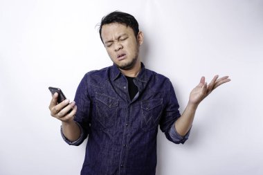 A thoughtful young Asian man is wearing blue shirt holding his phone and looks confused, isolated by white background