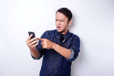 Shocked Asian man wearing blue shirt and holding his phone, isolated by white background