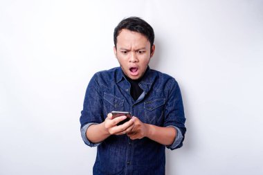 Shocked Asian man wearing blue shirt and holding his phone, isolated by white background