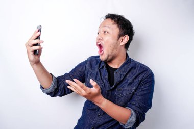 Shocked Asian man wearing blue shirt and holding his phone, isolated by white background