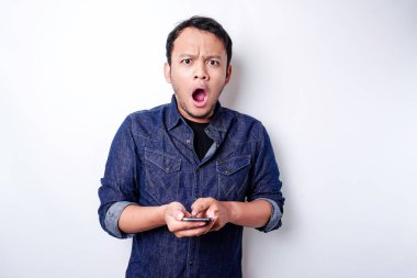 Shocked Asian man wearing blue shirt and holding his phone, isolated by white background