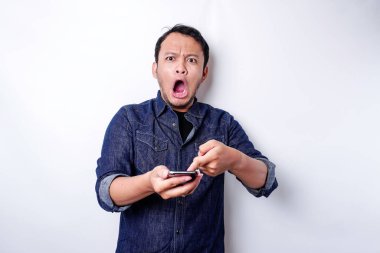 Shocked Asian man wearing blue shirt and holding his phone, isolated by white background