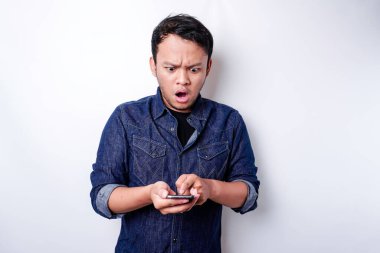 Shocked Asian man wearing blue shirt and holding his phone, isolated by white background