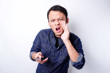 The angry and mad face of Asian man in blue shirt holding his phone isolated white background.