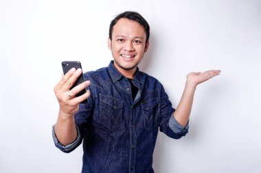 Excited Asian man wearing blue shirt pointing at the copy space beside him while holding his phone, isolated by white background