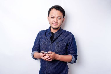 Excited Asian man wearing blue shirt smiling while holding his phone, isolated by white background