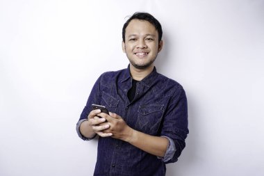 Excited Asian man wearing blue shirt smiling while holding his phone, isolated by white background