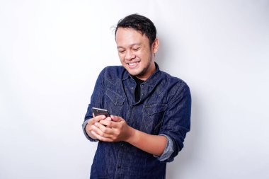 Excited Asian man wearing blue shirt smiling while holding his phone, isolated by white background