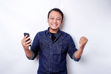 A young Asian man with a happy successful expression wearing blue shirt and holding his phone, isolated by white background
