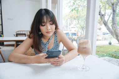 Sad face of a young Asian woman holding a cell phone wearing a tank top sitting near a window. Advertisement concept.