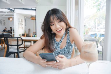 A happy Asian woman smiling in a restaurant, wearing a tank top and holding her phone