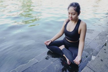 Asian woman practicing yoga and meditates near swimming pool outdoor. Fitness Asian lady sitting in lotus pose with closed eyes.