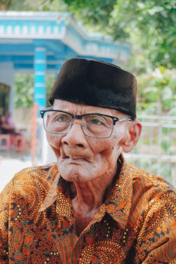 A portrait of an old Asian man wearing batik shirt and glasses