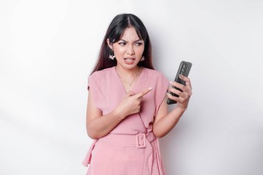 A dissatisfied young Asian woman dressed in pink, looks disgruntled with irritated face expressions holding her phone