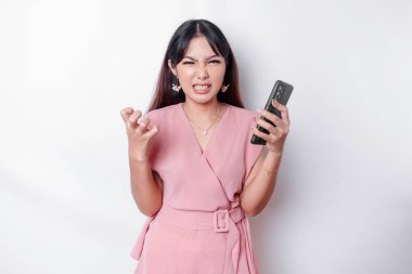 A dissatisfied young Asian woman dressed in pink, looks disgruntled with irritated face expressions holding her phone