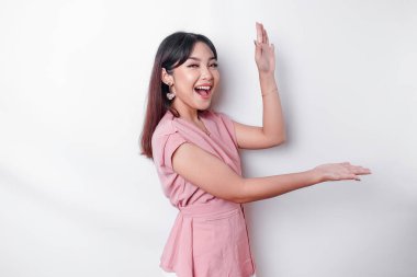 Excited Asian woman dressed in pink, pointing at the copy space beside her, isolated by white background
