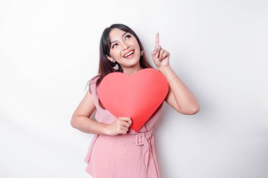 Portrait of a smiling Asian woman holding a big red heart symbol pointing up at copy space isolated over white background